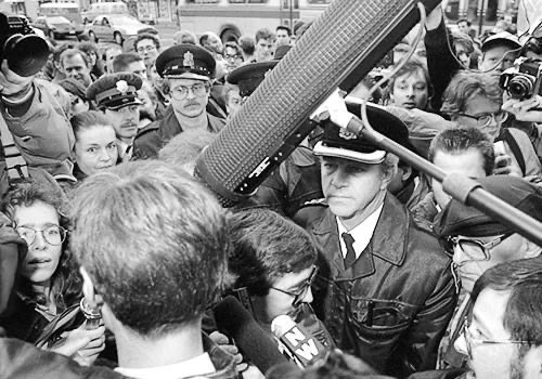 PICTURE: black and white photo of Gibson (foreground with back of head to the camera) encountering a large crowd of police, journalists and protesters.